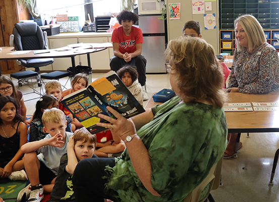 A woman in a chair holds up an open Dogman book to a class of kids. A woman watches from the side.